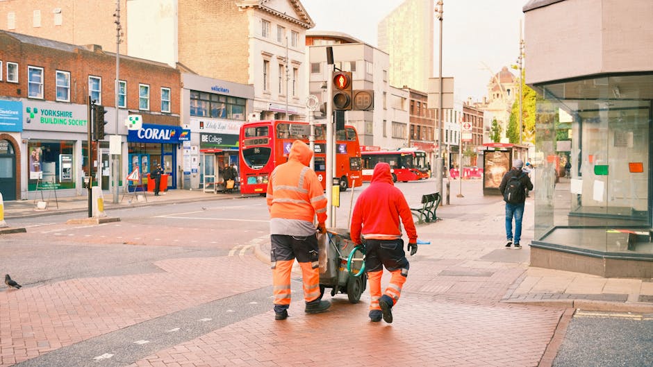 Two waste collection workers dressed in high-visibility orange and red jackets and trousers are walking across a busy city street, pushing a cart loaded with cleaning tools and waste containers. They 
