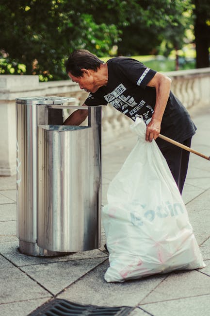 A wide alleyway between two urban buildings, cluttered with piles of mixed waste and discarded materials. In the foreground, a large, weathered grey bag with visible creases and stitched seams is posi
