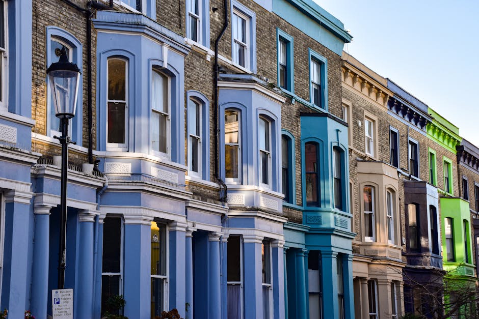 A busy street scene in Notting Hill, London, with rows of parked cars lining the road and a crowd of pedestrians walking along the pavement. The buildings on the left side of the street feature colorf