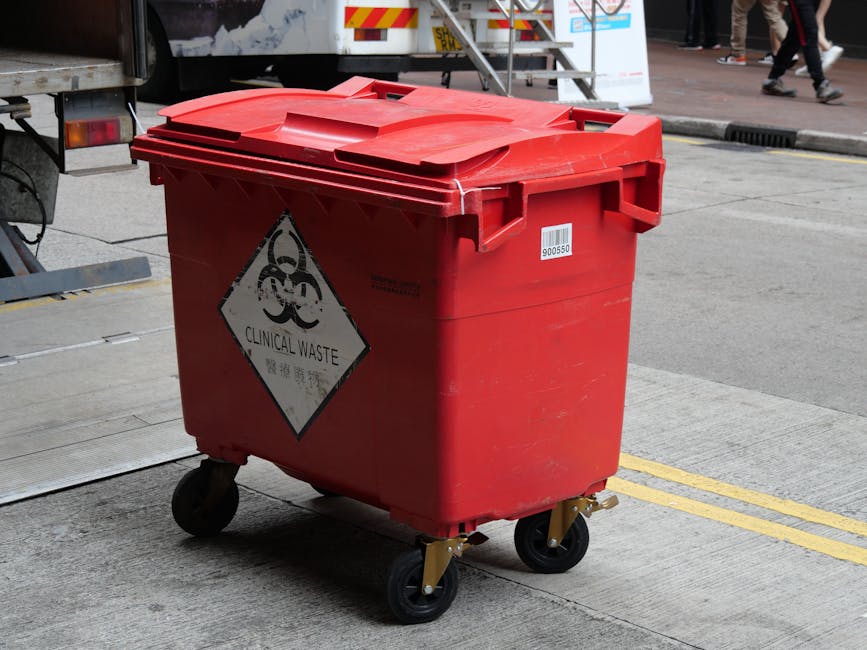 A large red plastic waste container with a closed lid, positioned on a concrete pavement in an urban outdoor setting. The container features a black and white hazardous waste symbol, including a bioha