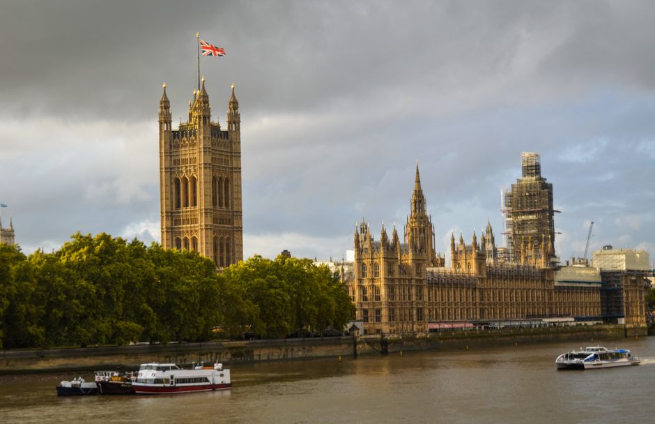 A panoramic view of the River Thames in London showing the historic Houses of Parliament and the Elizabeth Tower, commonly known as Big Ben, with a partly cloudy sky overhead. On the river, several bo